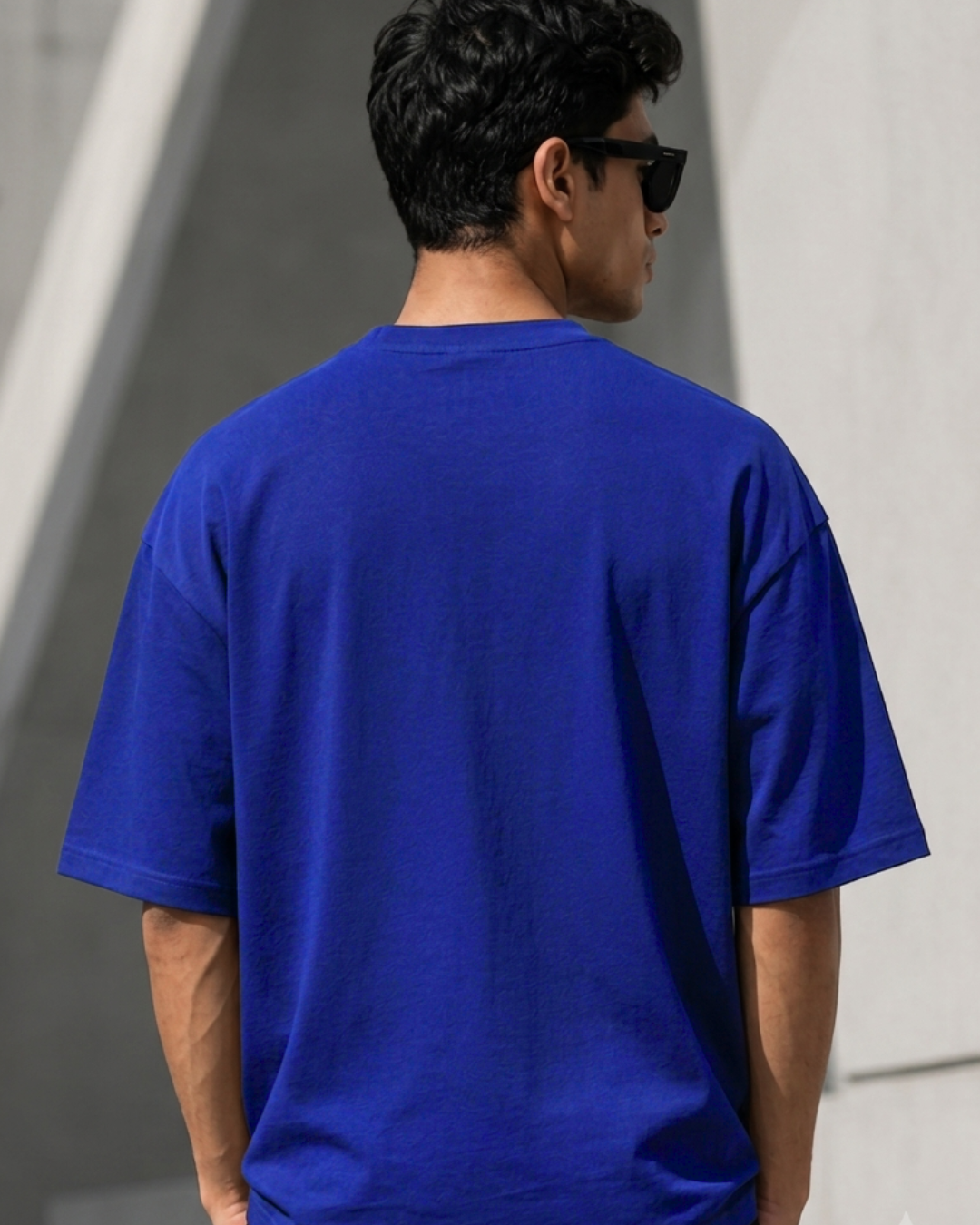 A man viewed from behind wearing a plain royal blue short-sleeve t-shirt and black sunglasses, standing against a light gray concrete wall.