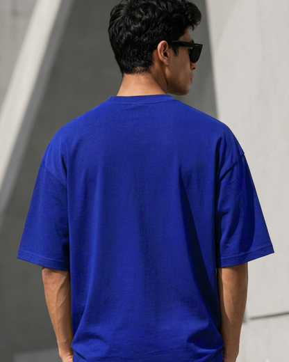 A man viewed from behind wearing a plain royal blue short-sleeve t-shirt and black sunglasses, standing against a light gray concrete wall.