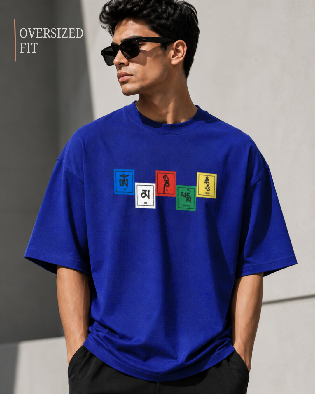 A man wearing an oversized royal blue t-shirt from white bear brand with a colorful graphic print on the chest, styled with black sunglasses and black pants against a gray concrete wall.