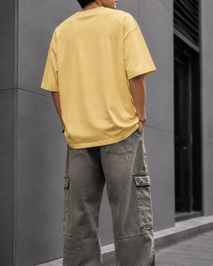 A man wearing an oversized beige graphic t-shirt with a distressed Union Jack design and a red stripe, paired with loose brown cargo pants, leaning against a gray wall.relaxed streetwear fit from white bear
