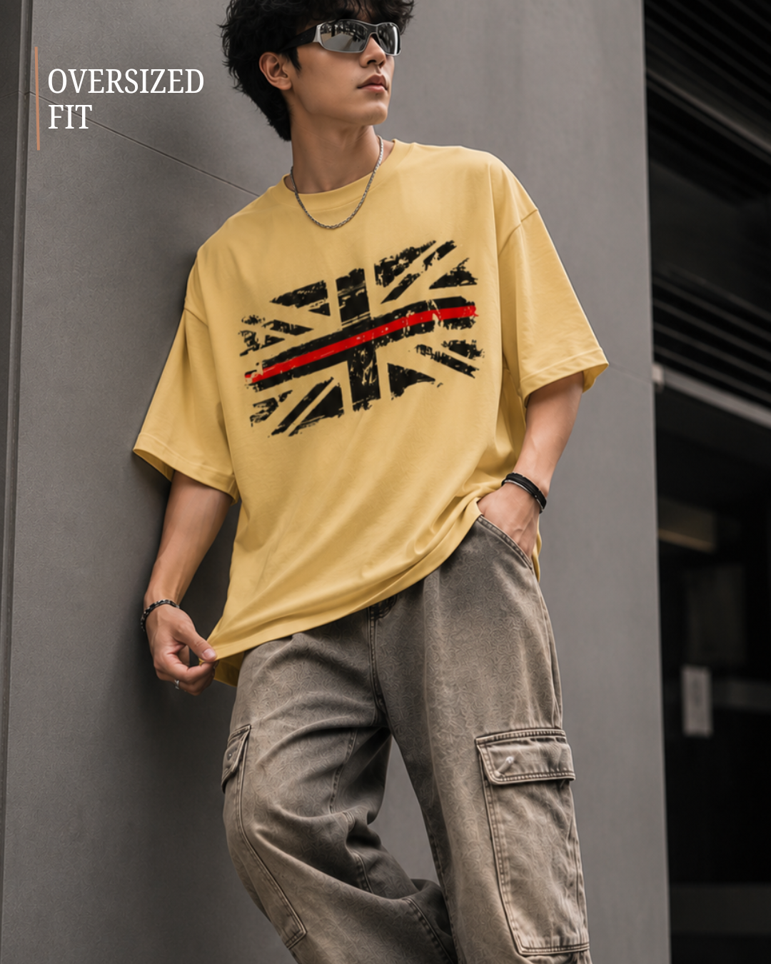A man wearing an oversized beige graphic t-shirt with a distressed Union Jack design and a red stripe, paired with loose brown cargo pants, leaning against a gray wall.relaxed streetwear fit from white bear