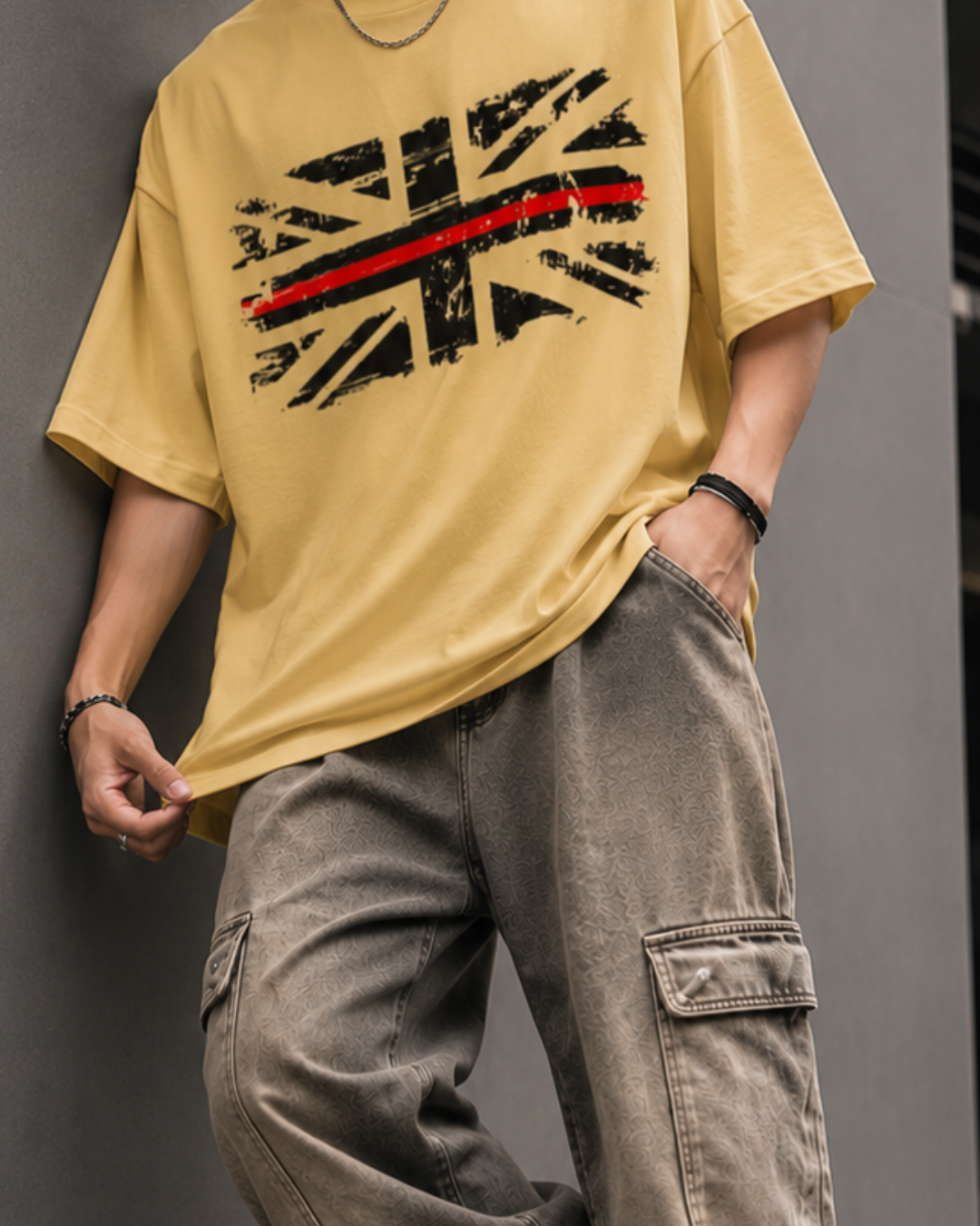 A man wearing an oversized beige graphic t-shirt with a distressed Union Jack design and a red stripe, paired with loose brown cargo pants, leaning against a gray wall.relaxed streetwear fit from white bear