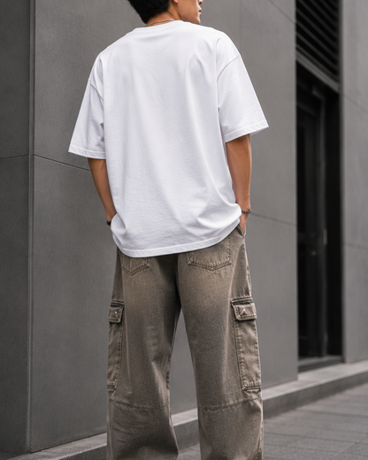 A man wearing an oversized white graphic t-shirt with a distressed Union Jack design and a red stripe, paired with loose brown cargo pants, leaning against a gray wall.relaxed streetwear fit from white bear