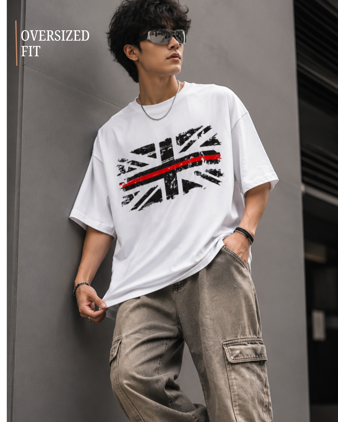 A man wearing an oversized white graphic t-shirt with a distressed Union Jack design and a red stripe, paired with loose brown cargo pants, leaning against a gray wall.relaxed streetwear fit from white bear