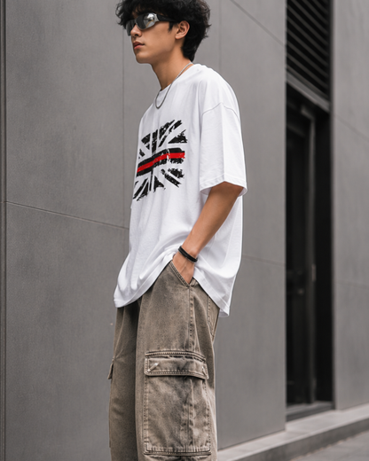 A man wearing an oversized white graphic t-shirt with a distressed Union Jack design and a red stripe, paired with loose brown cargo pants, leaning against a gray wall.relaxed streetwear fit from white bear