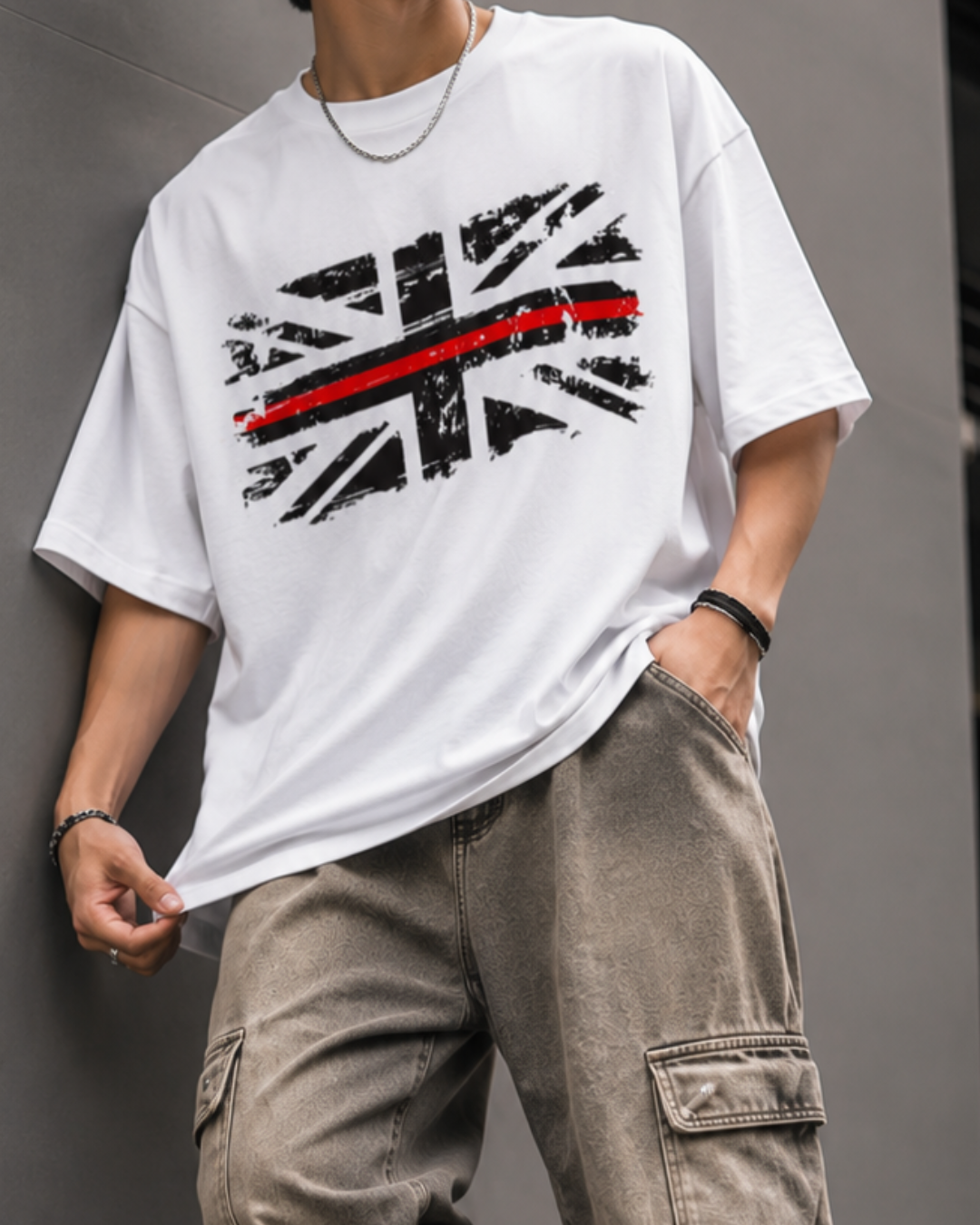 A man wearing an oversized white graphic t-shirt with a distressed Union Jack design and a red stripe, paired with loose brown cargo pants, leaning against a gray wall.relaxed streetwear fit from white bear